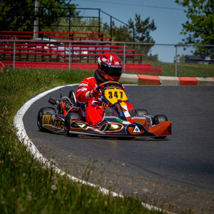 Piloto de kart en una curva, vestido de rojo, con fondo de gradas y vegetación.